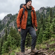 man wearing clothing layers including a jacket, beanie hat, tactical pants, boots in a forest