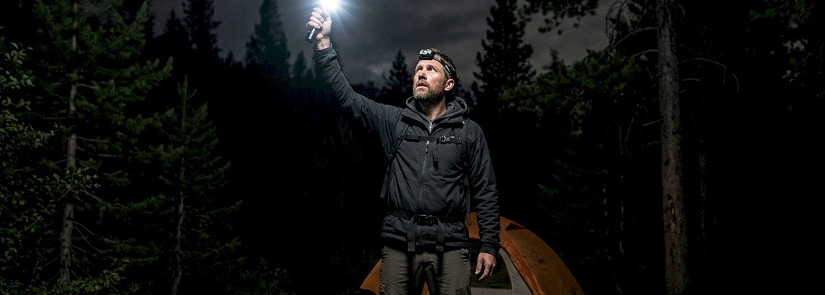 man in a forest holding large strobe flashlight at night near a camping site