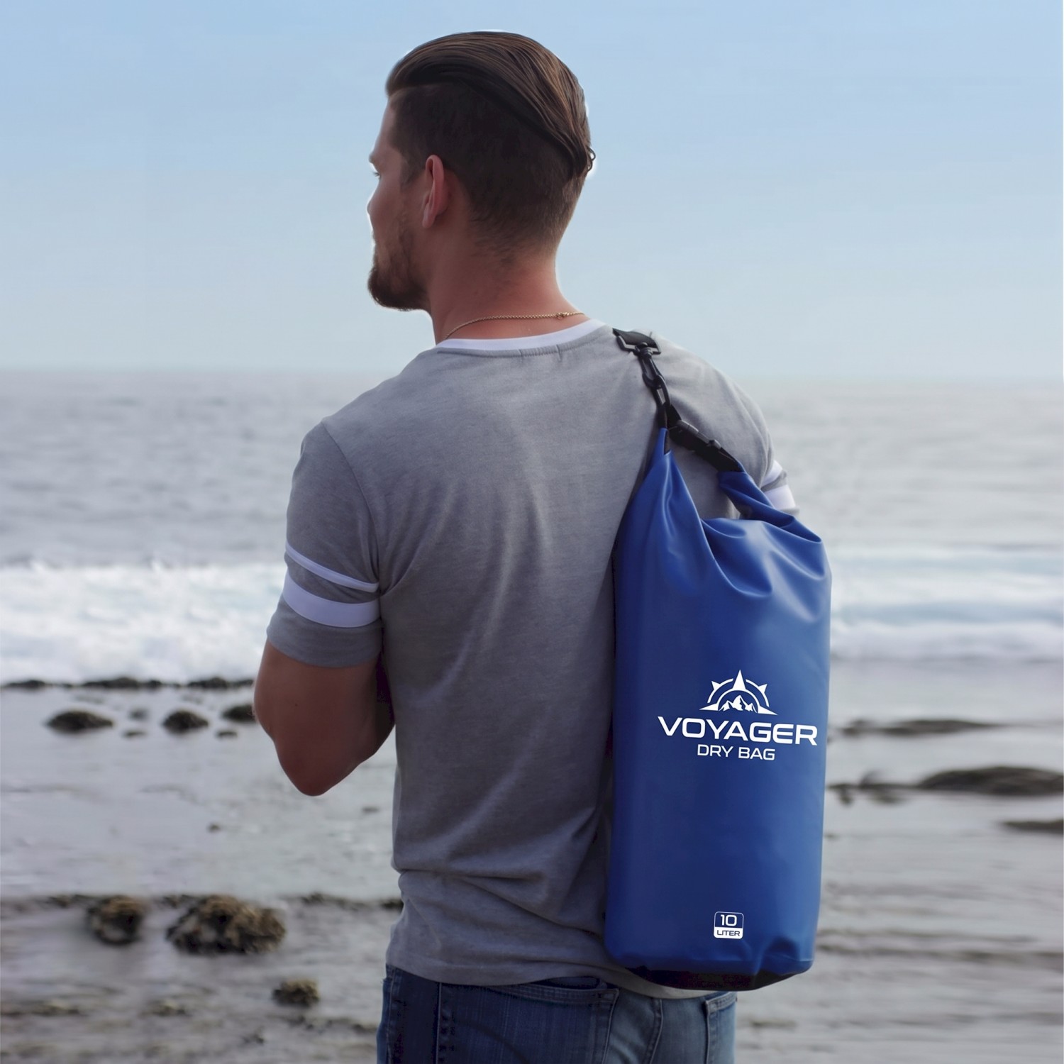 man holding a waterproof dry bag over his shoulder while standing on the beach looking over the Pacific Ocean