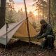 Backpacker setting up a rugged tactical tarp shelter in a forest clearing at sunrise