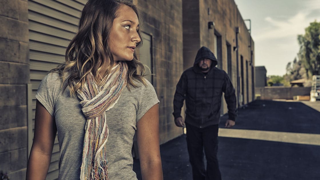 A woman needing self-defense in a dark alley, minimal illuminating light with a person following close behind